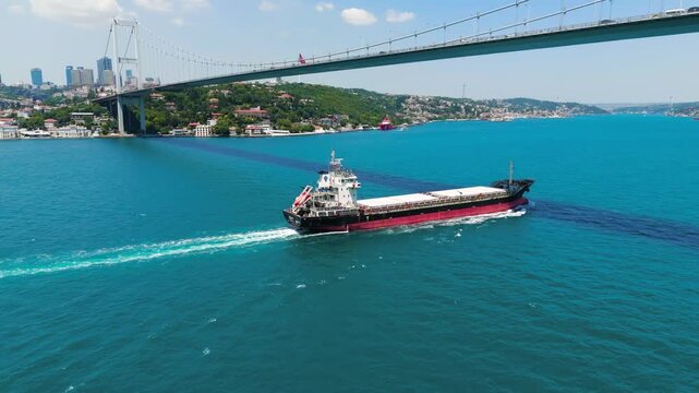 Istanbul, Turkey. 15 July Martyrs Bridge, a steel suspension span over the Bosphorus Strait, with a red-hulled bulk carrier sailing beneath on a bright sunny day, aerial view