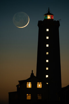 View of Grotta Lighthouse silhouetted against a twilight sky with a crescent moon and glowing windows in Seltjarnarnes, Seltjarnarnesbaer, Iceland.