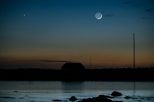 View of the crescent moon and a bright star shining in the twilight sky over a silhouetted building and calm water at Seltjarnarnes, Seltjarnarnesbaer, Iceland.