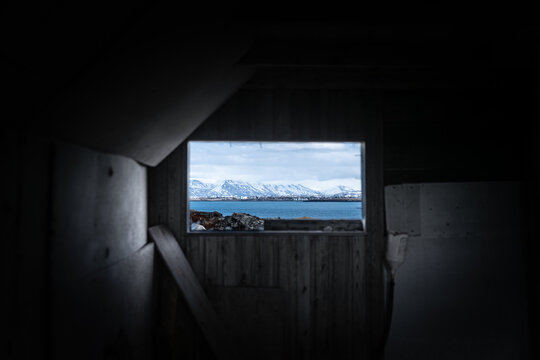 View of abandoned farmhouse interior looking out towards snowy mountains and the sea through a wooden window frame Hafnarfjordur, Hafnarfjardarkaupstad, Iceland.