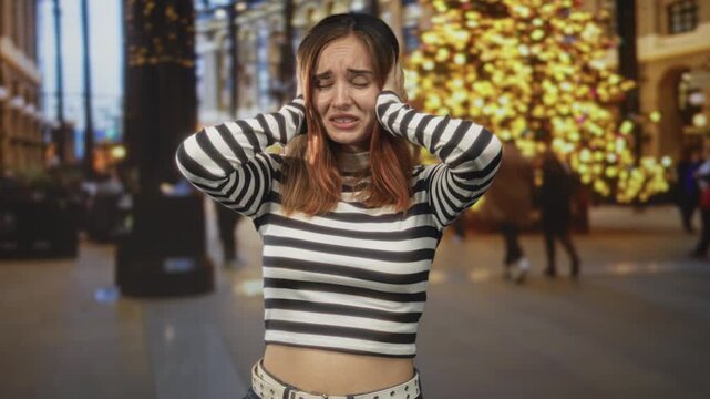 Young woman covering ears with hands in crowded building by large lit tree wearing striped crop top showing midriff; anxiety distress.