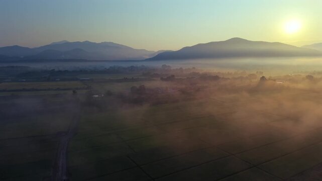 Cinematic aerial pull back shot of a foggy valley at sunrise Features a bright sun glowing over misty mountains and emerald farming plots in a quiet and majestic eastern regional district