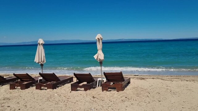 Empty wooden sun loungers on a beach on the Kassandra Peninsula in Chalkidiki, Greece, off season atmosphere as turquoise Mediterranean waves wash onto the sandy shore under bright spring light