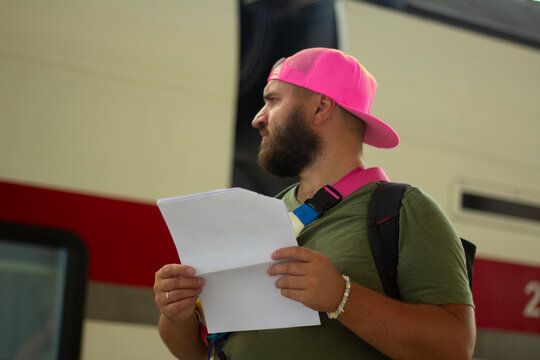 Horizontal photo. Young adult white man tourist with beard, pink baseball cap, printed ticket on paper sheet, stands on railway station platform looks at train. Concept of tourism, road trip, travel