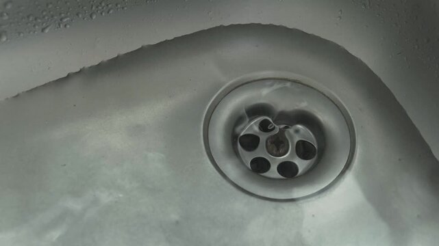 Hand pulls a sink plug. Water swirl down the drain of kitchen sink. Concept of domestic plumbing, household cleaning, and water drainage systems.