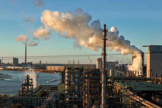 View of an industrial steel plant with smoking chimneys and pipes along the Rhine river under a clear sky at sunset in Duisburg, North Rhine-Westphalia, Germany.