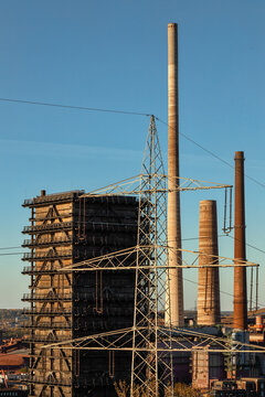 View of industrial chimneys and a high-voltage power pylon standing against a clear blue sky at an old steel mill site in Duisburg, North Rhine-Westphalia, Germany.