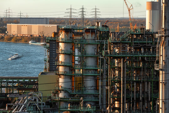 View of an industrial plant with steel towers and complex piping near a river with cargo ships at sunset in Duisburg, North Rhine-Westphalia, Germany.