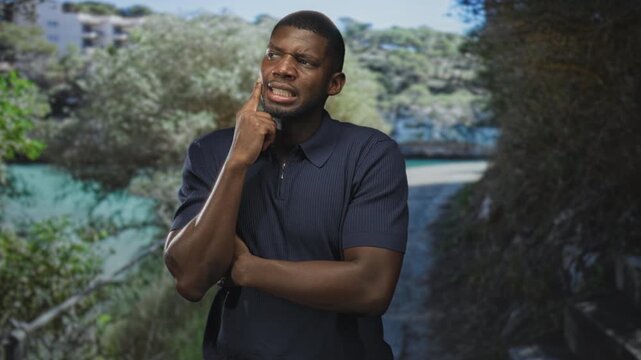 Man touching cheek with finger, arms crossed on a street path beside water and trees, pensive expression; thoughtful doubt.