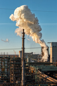 View of a large industrial complex with a massive plume of white smoke rising from a tall chimney stack under a clear blue sky during sunset Duisburg, North Rhine-Westphalia, Germany.