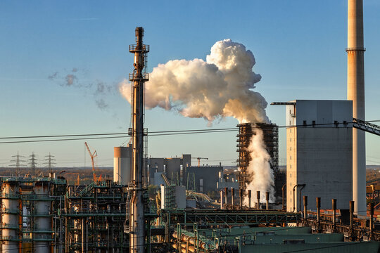 View of an industrial plant with thick white smoke rising from a chimney stack against a clear blue sky during sunset in Duisburg, North Rhine-Westphalia, Germany.