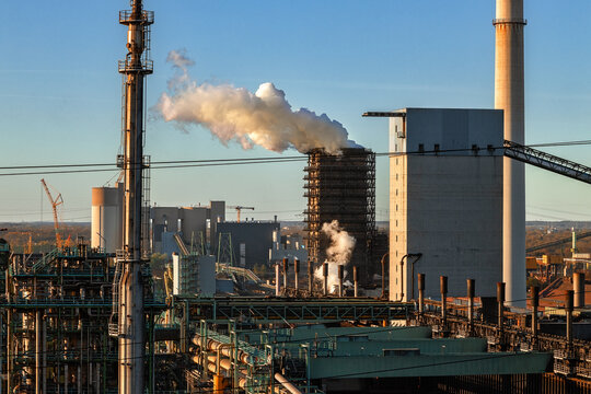 View of an industrial steel plant with smoking chimneys and complex piping under a clear blue sky during golden hour in Duisburg, North Rhine-Westphalia, Germany.