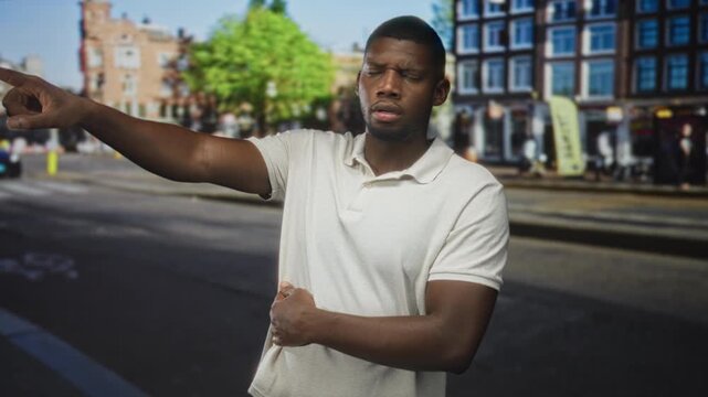 Young man points with outstretched arm while clutching stomach and showing stomachache on a busy urban street lined with buildings and cafes; concern urgency.