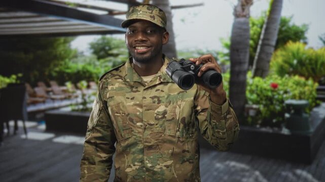 Soldier man holding binoculars, smiling and looking up while standing on a building terrace with palm trees; pride duty vigilance.
