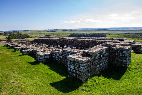 View of the ancient Roman stone ruins of Vindolanda fort with green grass and rolling hills under a bright sky Hexham, England, United Kingdom.