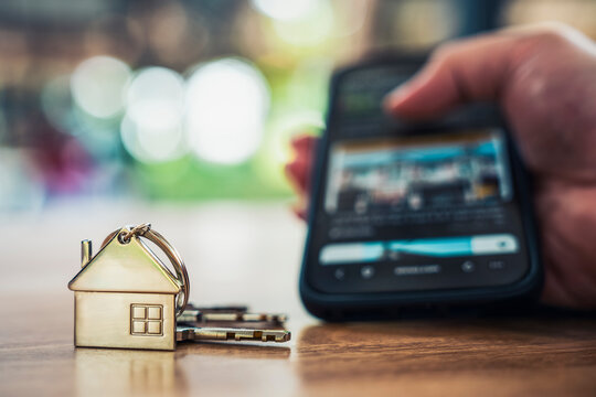 House model keys on wooden table with blurred person using smartphone in background. Online real estate search, property investment, smart home control and buying house concept.