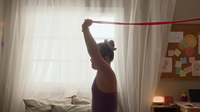 Tilt down shot of focused teenage Caucasian girl with Down syndrome wearing lilac leotard and white tights standing in cozy room and rotating gymnastics rope with outstretched arm during home workout