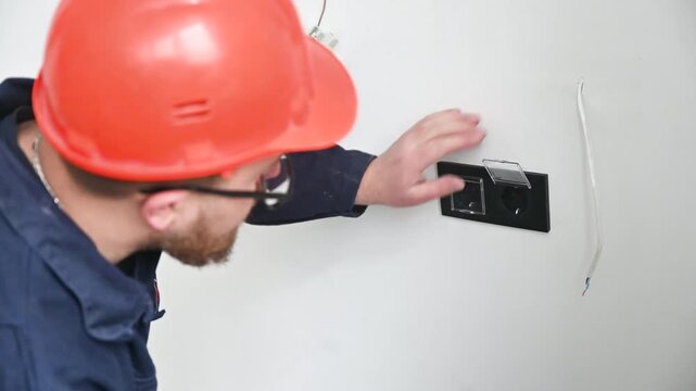 Electrician repairs and installs power outlet in residential room. Worker adjusts socket frame and cable connection during interior construction and setup