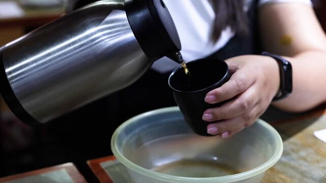 Woman pouring hot tea from silver thermos into black cup at restaurant table.