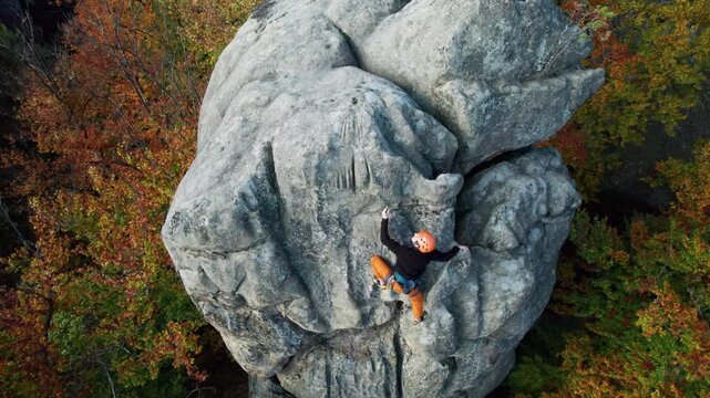 Man climbs natural sandstone face in autumn woodland, wearing harness and safety gear. Drone footage for outdoor training, strength, focus, and extreme sport storytelling