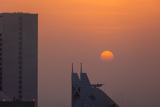 View of the sun setting behind modern skyscrapers and architectural structures under a hazy orange sky Dubai, Dubai, United Arab Emirates.