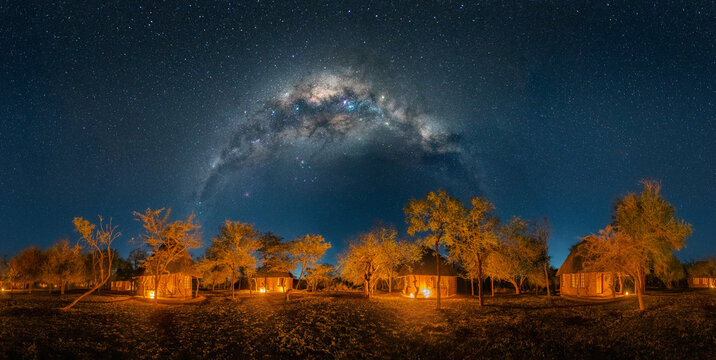 View of Ndlovu Camp in Hlane Royal National Park featuring illuminated traditional huts under a vibrant Milky Way arch and starry night sky in Simunye, Lubombo Region, Eswatini.