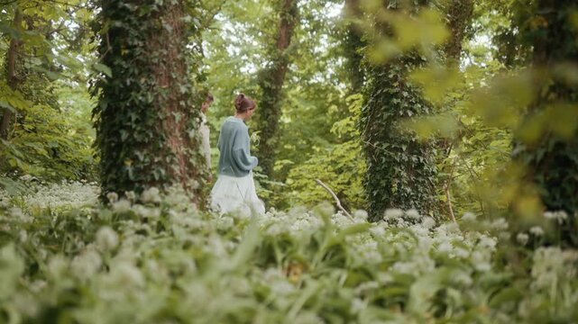 A couple walks through a lush green woodland path filled with wild garlic flowers.