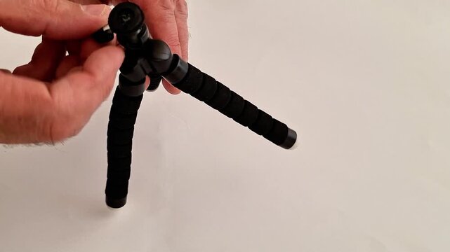 Hand holding and adjusting a compact black tripod with flexible textured legs and camera mount, shown against a plain white background, highlighting portable photography gear