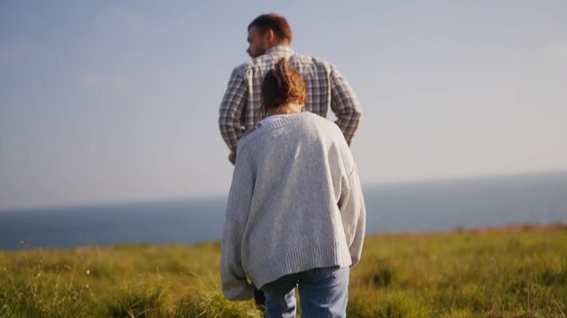 A father and daughter look out over the expansive blue ocean from a grassy cliff.