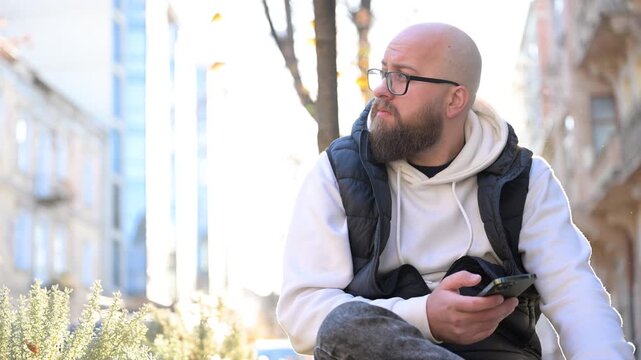Bearded man dressed in casual attire sits on bench along bustling city street, checking phone while enjoying warm morning sunlight. Surrounding buildings create urban backdrop full of life.