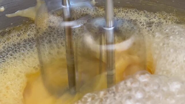A side view macro video shows the whisk of an automatic stand mixer rotating through eggs to create thick, fluffy white foam and bubbles in a stainless steel bowl during baking.