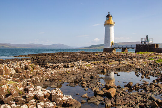 View of the Tobermory Lighthouse standing on a rocky shoreline with distant hills and a clear blue sky Isle of Mull, Scotland, United Kingdom.