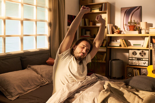 Young adult Caucasian man stretching in bed after waking, starting morning routine at home and expressing rest, recovery, work life balance. Lifestyle and wellness marketing use