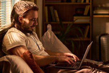 Young adult Caucasian man working on laptop while sitting on bed at home, managing remote tasks in...