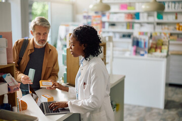 Fototapeta premium Black female pharmacist using laptop while talking to customer in drugstore.