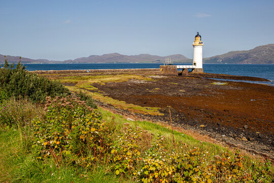 View of Tobermory Lighthouse standing on the rocky coastline surrounded by green vegetation and the blue waters of the Sound of Mull with distant hills Isle of Mull, Scotland, United Kingdom.