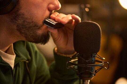 Close up of young adult man playing harmonica into studio microphone while recording audio content. Suitable for music production, podcast branding, live session promotion