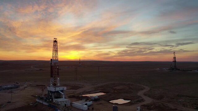 Oil drilling rig standing against dramatic sunset sky in desert field.