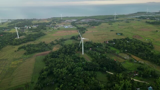 Aerial orbit of wind turbines across coastal farmland with green fields and distant shoreline in Guimaras, Visayas, Philippines.