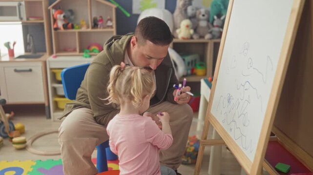 Man guiding girl drawing on whiteboard, holds marker and points to scribbles in a playroom building; tender learning bonding.