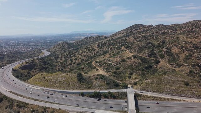 Multi lane highway with steady traffic passing through dry hills in Simi Valley California under clear blue sky with winding dirt trails rugged terrain and suburban landscape. Aerial panoramic shot.
