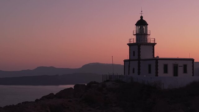 Drone orbiting the Santorini lighthouse at sunrise, capturing a serene atmosphere with calm light, caldera cliffs, and the peaceful Aegean Sea.