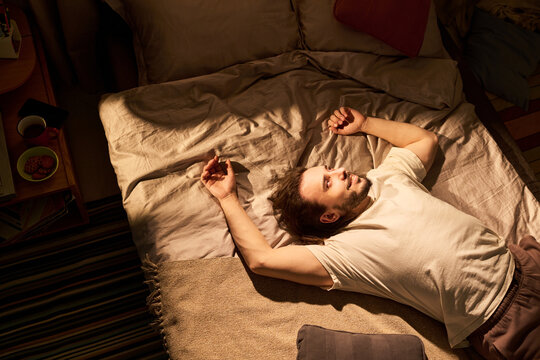 High angle view of Caucasian young adult man lying awake on bed, struggling with insomnia and stress at home. Useful for sleep disorder, mental health, burnout campaigns