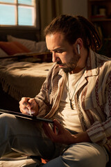 Obraz premium Vertical shot of young adult man using tablet while wearing wireless earbuds at home, browsing digital content. Suitable for remote lifestyle, streaming service, or mobile app marketing