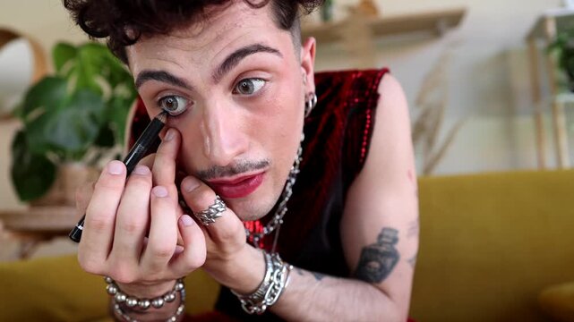 Young gender nonconforming man with tattoos and jewelry carefully applying black eyeliner pencil to his lower lash line while sitting on a sofa, challenging traditional gender norms