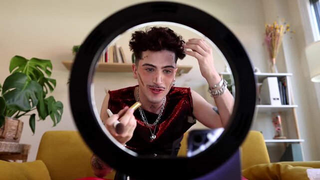 Young male content creator with an extravagant style applying contouring and foundation with a brush while filming a makeup tutorial for social media with a ring light and smartphone