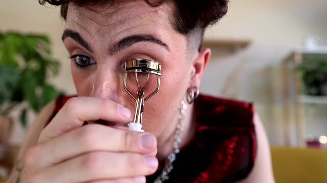 Young non binary person with mustache and makeup meticulously curling their eyelashes with a golden eyelash curler as part of their daily beauty routine in a bright, modern living room
