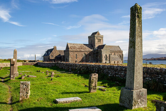 View of Iona Abbey and Nunnery featuring historic stone architecture, an ancient graveyard with obelisks, and a low stone wall under a bright blue sky Isle of Iona, Scotland, United Kingdom.