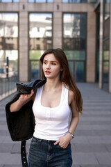 Fototapeta premium A beautiful brunette in a leather jacket, jeans and sneakers walks down the street of a modern city.