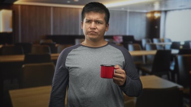 Man holding red mug near chest in a cafeteria building, wearing grey raglan shirt and squinting with a displeased look; morning skepticism.
